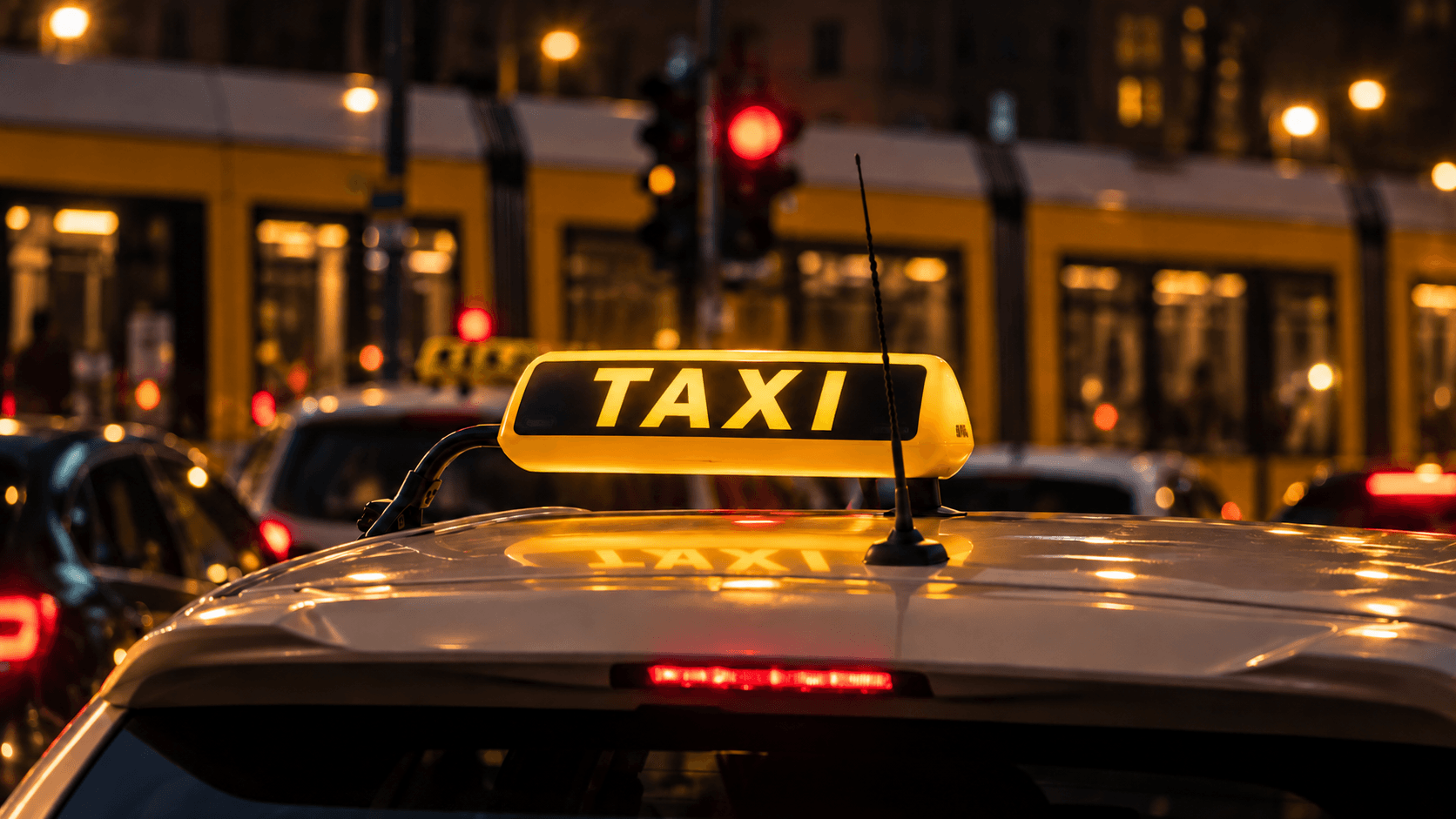 Apšviestas geltonas TAKSI ženklas ant automobilio stogo naktiniame mieste